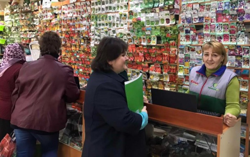 На малюнку зображено крамницю з насінневим матеріалом в пакетиках, продавця і фахівця Держпродспоживслужби