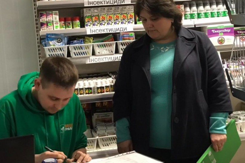На малюнку зображено крамницю з насінневим матеріалом в пакетиках, продавця і фахівця Держпродспоживслужби