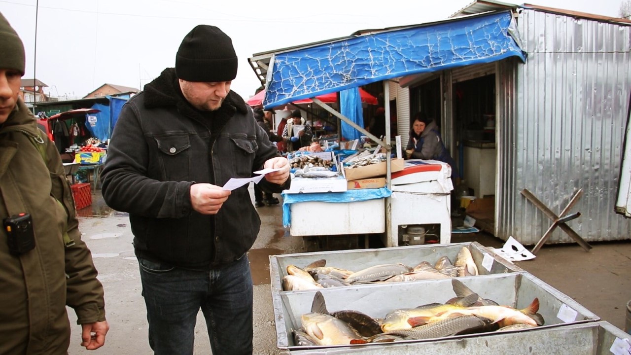 На малюнку зображено двох чоловіків на ринку біля коробів з живою рибою